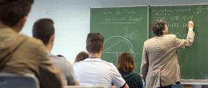 Ein Mathematiklehrer steht mit dem Rücken zur Klasse und schreibt mit Kreide Mathematikaufgaben an eine Tafel.