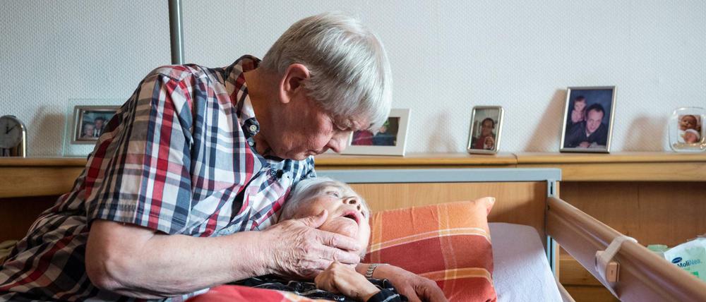 Ein pflegender Ehemann nimmt in seiner Wohnung seine an Demenz und Parkinson erkrankte Ehefrau in den Arm.