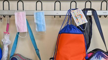 Masken hängen zusammen mit Taschen und Rucksäcken an Kleiderhaken in einem Klassenraum einer Grundschule.