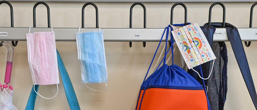 Masken hängen zusammen mit Taschen und Rucksäcken an Kleiderhaken in einem Klassenraum einer Grundschule.