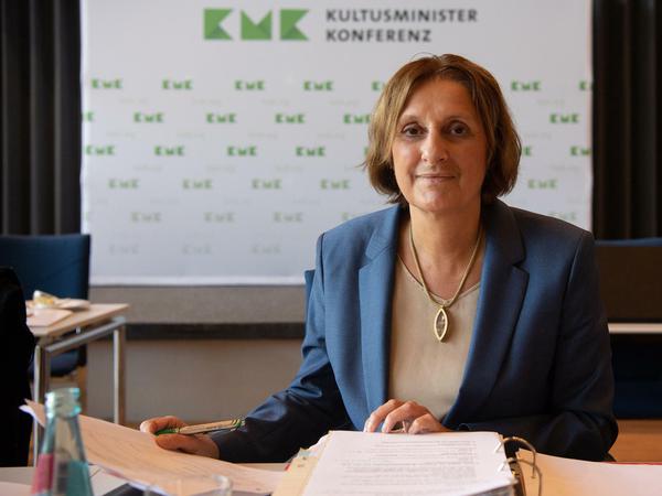 Ein Porträtfoto von Britta Ernst an ihrem Schreibtisch, im Hintergrund ist das Logo der KMK zu sehen.