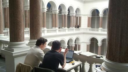 Im Kreuzgang des Lichthofs der TU Berlin sitzen zwei Studierende vor ihren Laptops nebeneinander an einem Tisch.