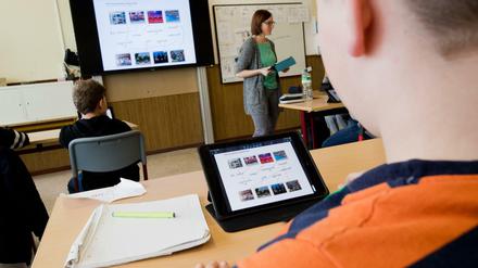 Ein Schüber sitzt vor einem Programmmenü auf seinem IPad in der Klasse, vorne an der digitalen Tafel erscheint dasselbe Bild.