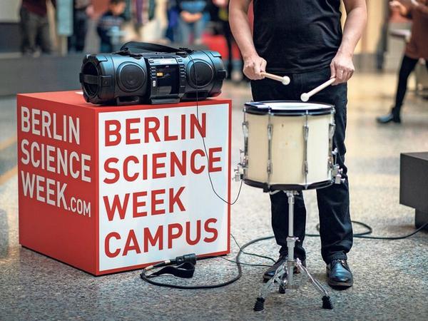 Neben einem Kubus mit der Aufschrift Berlin Science Week Campus steht ein Trommler mit Instrument.