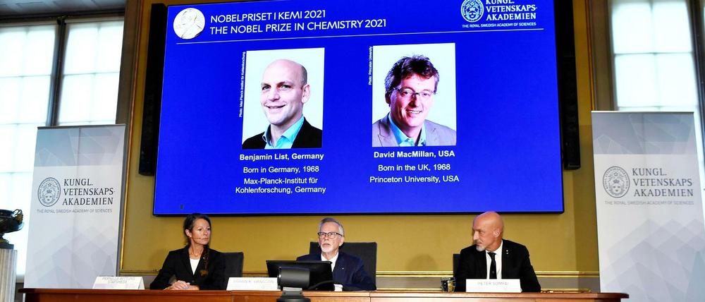An einem Podiumstisch sitzen zwei Männer und eine Frau, über ihnen die Bilder von zwei Nobelpreisträgern.