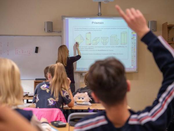 Schüler sitzen in einer Klasse, in der die Lehrerin vorne am Smartboard steht.