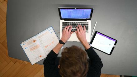 Ein Lehrer sitzt mit einem Schulbuch links und einem Tablet rechts an seinem Platz im Lehrerzimmer.