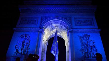 Menschen versammeln sich vor dem blau beleuchteten Arc de Triomphe in Paris, in dessen Bogen eine EU-Flagge hängt