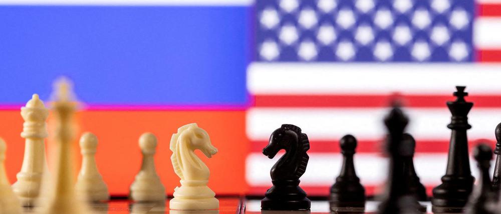 Das Bild zeigt Schachfiguren und im Hintergrund die Fahnen Russlands und der USA. 