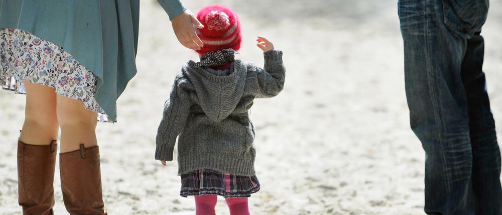 Ein Kleinkind steht mit zwei Erwachsenen auf einem Spielplatz im Sand.