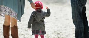 Ein Kleinkind steht mit zwei Erwachsenen auf einem Spielplatz im Sand.