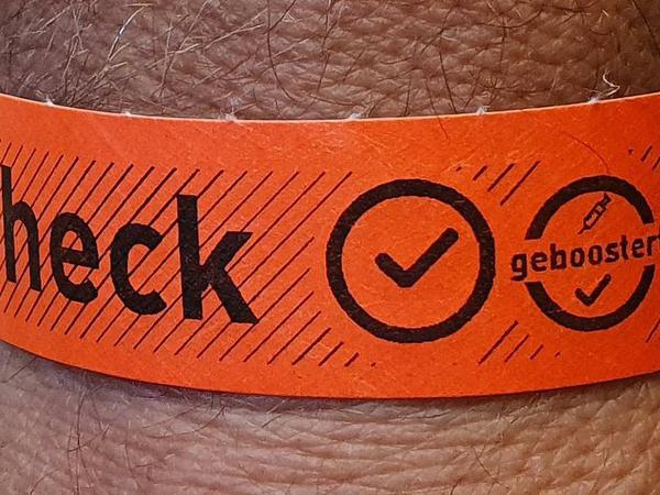 Ein Handgelenk ist von einem orangenen Papierarmband umschlossen, auf dem "Check", ein Häkchen und ein Symbol mit der Aufschrift "geboostert" zu lesen sind.