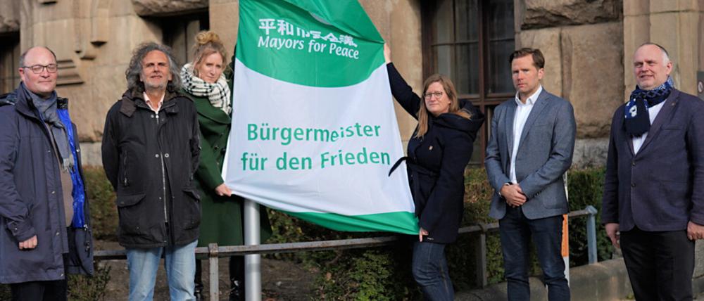 Bürgermeisterin Kirstin Bauch und die Stadträt:innen Arne Herz, Oliver Schruoffeneger, Heike Schmitt-Schmelz, Fabian Schmitz-Grethlein und Detlef Wagner hissen "Mayors for Peace"-Flagge.