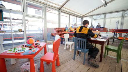 Ein größerer Junge sitzt in einem Zelt mit Plexiglaswänden an einem Tisch und hantiert mit Stiften und Papier.