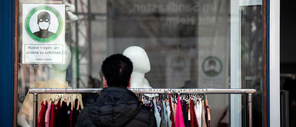 Eine Frau schaut sich Kleidung an einer Stange vor einem Geschäft an, auf einem Schild im Fenster steht "Maske tragen, um andere zu schützen".