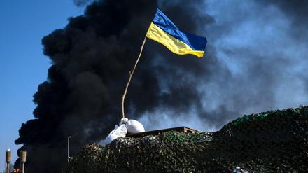 Kiew: Eine ukrainische Flagge weht auf einem Kontrollpunkt, während schwarzer Rauch aus einem Treibstofflager der ukrainischen Armee nach einem russischen Angriff aufsteigt.