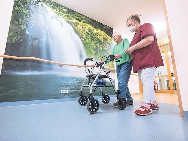 Eine Pflegerin begleitet eine weißhaarige Frau, die im Pflegeheim am Rollator geht. An der Wand ein Panoramabild von einem Wasserfall.