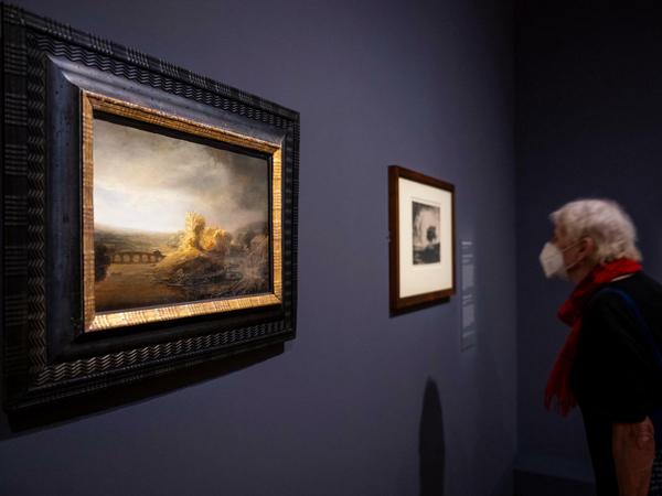 Das Landschaftsbild Rembrandts ist mit einem anderen Werk an einer Wand in einem Museumsraum zu sehen, im Bild auch eine Betrachterin.
