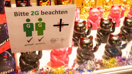 In einem Schaufenster in einem Ladengeschäft hängt ein Schild mit der Aufschrift „Bitte 2G beachten“. Im Hintergrund stehen Winkekatzen, die den Mittelfinger zeigen. 