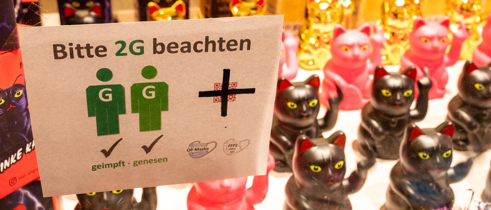 In einem Schaufenster in einem Ladengeschäft hängt ein Schild mit der Aufschrift „Bitte 2G beachten“. Im Hintergrund stehen Winkekatzen, die den Mittelfinger zeigen.