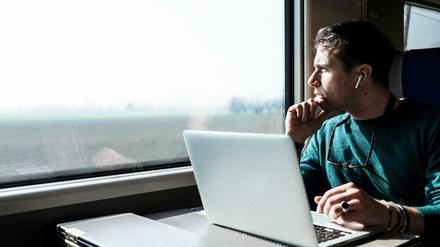 Ein junger Mann sitzt im Zug an einem Laptop und schaut nachdenklich aus dem Fenster.