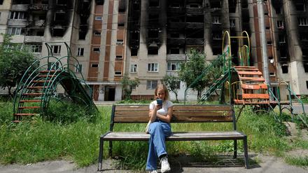 Ein Mädchen schaut in der ukrainischen Stadt Irpin auf sein Smartphone. Im Hintergrund ein stark beschädigtes Wohnhaus.
