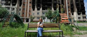Ein Mädchen schaut in der ukrainischen Stadt Irpin auf sein Smartphone. Im Hintergrund ein stark beschädigtes Wohnhaus.