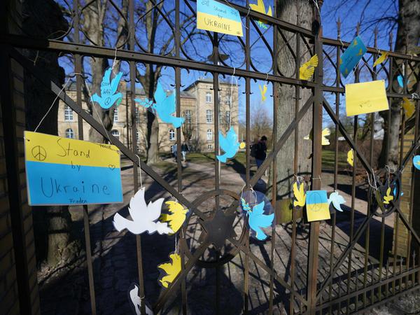 Hinter einem schmiedeeisernen Tor mit Ukraineflaggen und Friedenstauben in gelb-blau ist ein Schulgebäude zu sehen.
