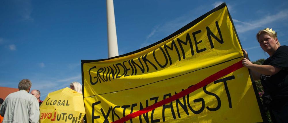 Teilnehmer einer Demonstration für ein bedingungsloses Grundeinkommen vor dem Fernsehturm mit einem Transparent mit dem Aufdruck Grundeinkommen.