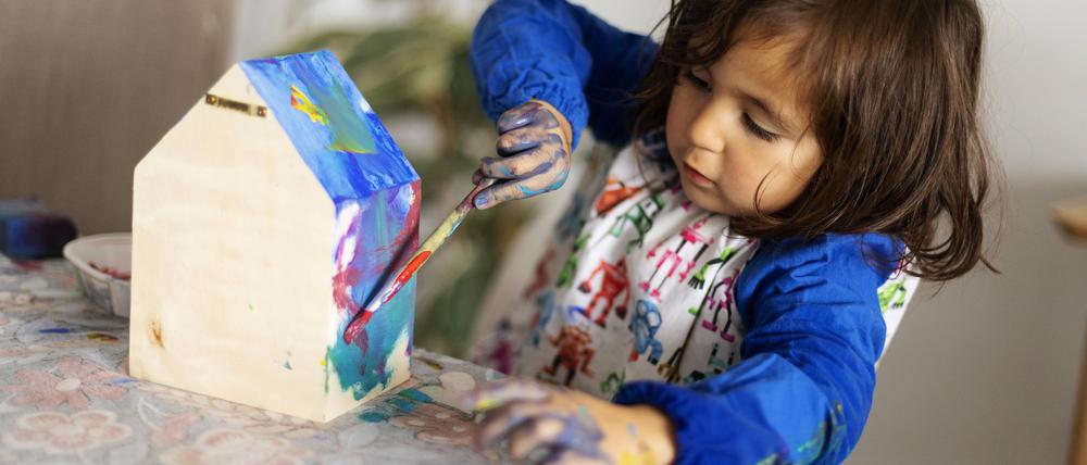 Cute girl painting small wooden model house with paintbrush in living room.