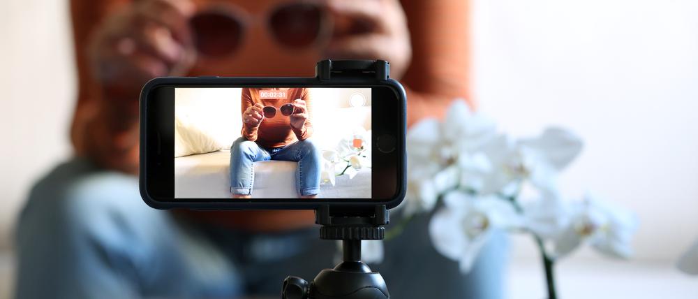 Eine Frau sitzt mit einer Sonnenbrille in Händen auf einem Sofa und filmt sich dabei mit ihrem iPhone (gestellte Szene). Produkttipps auf Instagram oder Tiktok sind oft bezahlte Werbung.
