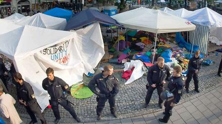Polizisten in schwerer Einsatzkleidung riegeln das bereits geräumte Camp in München ab, im Hintergrund sind Zelte und Berge von Isomatten und Schlafsäcken zu sehen.