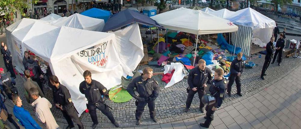 Polizisten in schwerer Einsatzkleidung riegeln das bereits geräumte Camp in München ab, im Hintergrund sind Zelte und Berge von Isomatten und Schlafsäcken zu sehen.