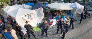 Polizisten in schwerer Einsatzkleidung riegeln das bereits geräumte Camp in München ab, im Hintergrund sind Zelte und Berge von Isomatten und Schlafsäcken zu sehen.