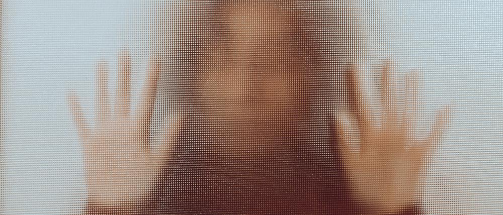Back lit image of the silhouette of a woman with her hands pressed against a glass window. The silhouette is distorted, and the arms elongated, giving an alien-like quality. The image is sinister and foreboding, with an element of horror. It is as if the 'woman' is trying to escape from behind the glass.