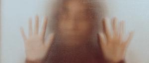 Back lit image of the silhouette of a woman with her hands pressed against a glass window. The silhouette is distorted, and the arms elongated, giving an alien-like quality. The image is sinister and foreboding, with an element of horror. It is as if the 'woman' is trying to escape from behind the glass.