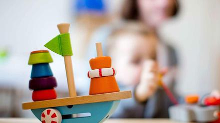 Auf einem Tisch in einem Kindergarten steht ein Holzspielzeug auf einem Tisch, im Hintergrund ist ein Kleinkind zu sehen.