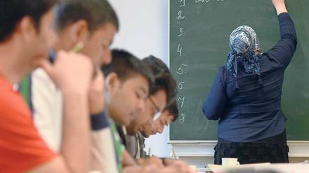 Eine junge Frau schreibt in einem Klassenraum etwas an die Tafel, junge Männer sitzen an Tischen und schreiben in ihre Hefte.