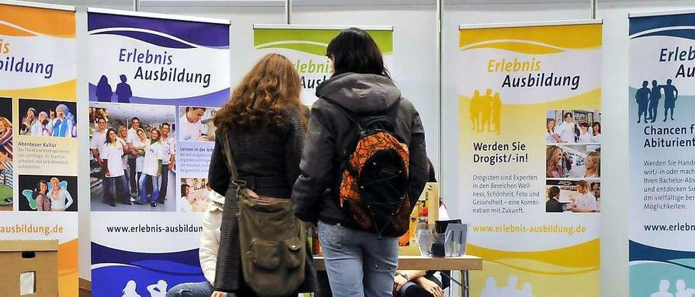 Zwei junge Frauen stehen vor einer Wand mit Plakaten, die auf einer Ausbildungsmesse für Berufe werben.