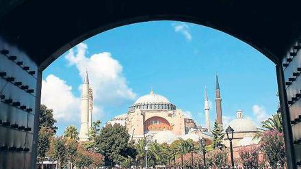 Menschen drängen sich auf einer Grünfläche, im Hintergrund ist eine große Moschee zu sehen. 