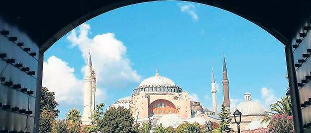 Menschen drängen sich auf einer Grünfläche, im Hintergrund ist eine große Moschee zu sehen. 