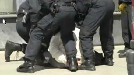 Das Video zeigt mehrere Beamte, die einen Mann in Handschellen am Boden halten. Der Beamte am linken Bildrand tritt darin mehrfach zu.