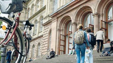 Studierende betreten ein Universitätsgebäude, einige sitzen auf der Treppe.