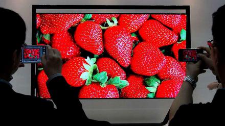 Auf einem rieseigen Bildschirm sind strahlend rote Erdbeeren zu sehen. Davor zwei Betrachter, die den Fernsehbildschirm fotografieren.