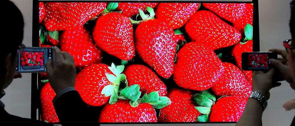 Auf einem rieseigen Bildschirm sind strahlend rote Erdbeeren zu sehen. Davor zwei Betrachter, die den Fernsehbildschirm fotografieren.