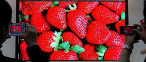 Auf einem rieseigen Bildschirm sind strahlend rote Erdbeeren zu sehen. Davor zwei Betrachter, die den Fernsehbildschirm fotografieren.