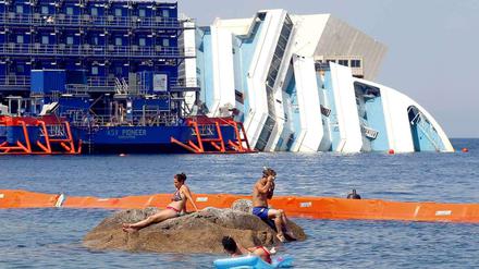 Was stört die Urlauber schon ein gesunkenes Kreuzfahrtschiff? Das Wrack der havarierten Costa Concordia vor der kleinen italienischen Insel Giglio. Die Bergung soll im September beginnen, doch offenbar hat sich das Schiff so stark verformt, dass das schwierig werden könnte. 