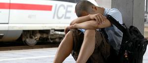 Ein junger Mann in kurzer Hose sitzt auf einem Bahnsteig, den Kopf auf die Arme gestützt. Im Hintergrund steht ein ICE-Zug.