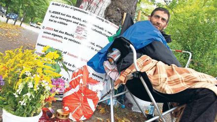 Unterm Baum. Firas Maraghy trinkt seit einem Monat nur Wasser. Er streikt für ein Reisedokument für seine Tochter. Unterstützer bringen ihm Blumen und Kerzen.