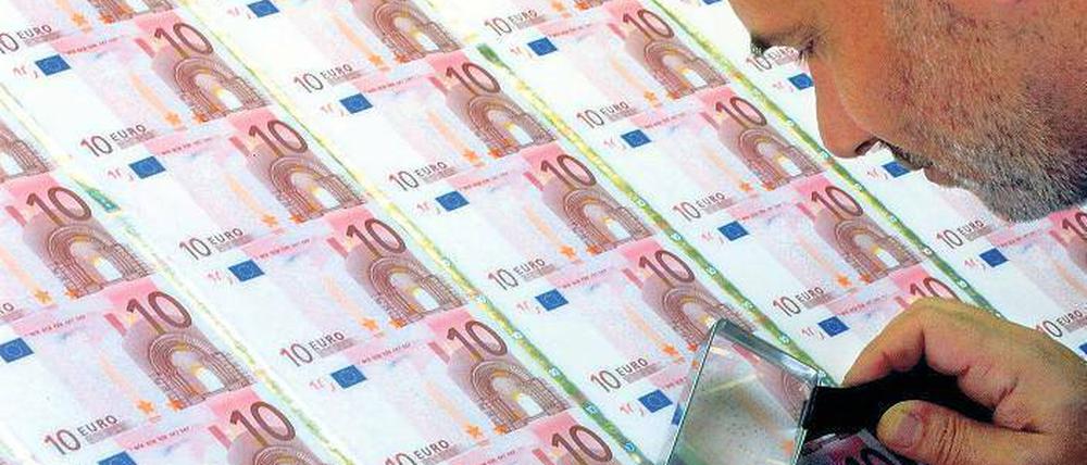 Ein Mann beugt sich mit einer Lupe in der Hand über einen Bogen mit Zehn-Euro-Geldscheinen.